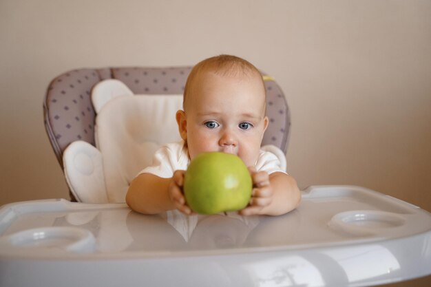 un enfant sur une chaise d'enfant dans un body blanc joue avec une pomme verte