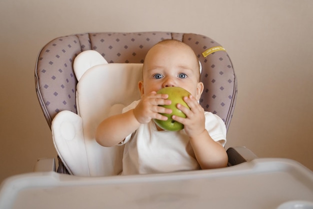 un enfant sur une chaise d'enfant dans un body blanc joue avec une pomme verte