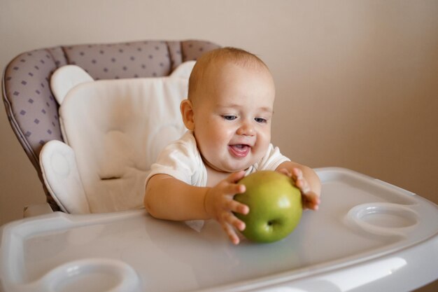 un enfant sur une chaise d'enfant dans un body blanc joue avec une pomme verte