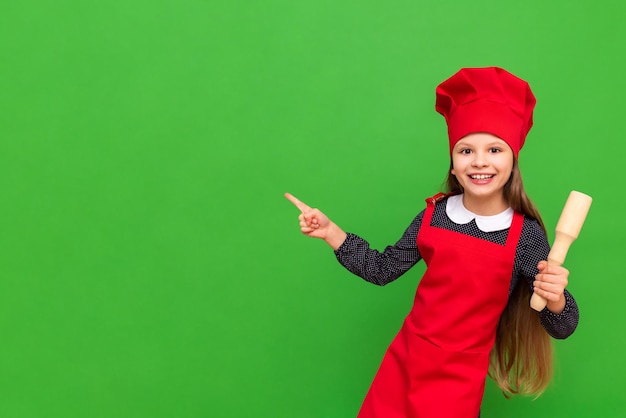 L'enfant chef pointe vers votre publicité sur un fond vert isolé Une petite fille vêtue d'un tablier rouge et d'une casquette tient une corde à sauter pour la pâte Espace de copie