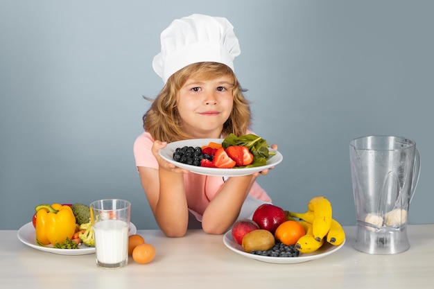 Un enfant chef tient une assiette avec des fruits isolés sur bleu Drôle de petit enfant chef cuisinier portant une casquette de cuisinier uniforme et un tablier des aliments cuits dans la cuisine