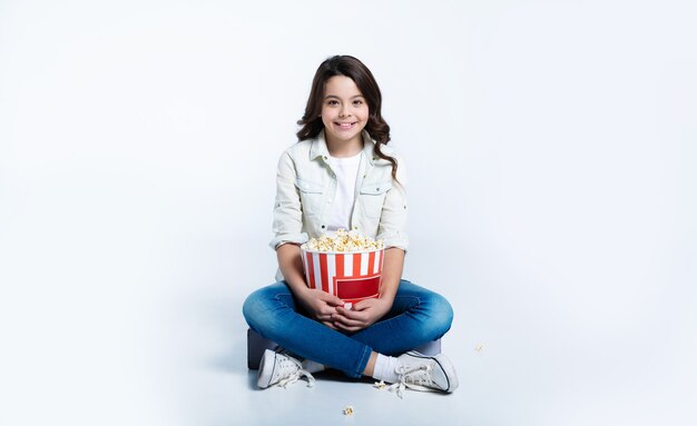 L'enfant concentré est assis dans une position de lotus et mange du pop-corn, tout en regardant des dessins animés ou des films.