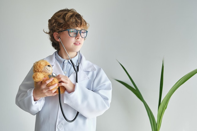 Enfant contemplatif en lunettes et robe médicale écoutant le battement du cœur d'un ours doux tout en détournant le regard sur un fond blanc