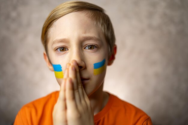 Un enfant contre la guerre un garçon avec un drapeau peint de l'Ukraine sur ses joues a joint ses mains dans un plaidoyer pour la paix mondiale