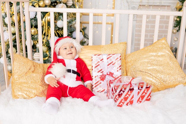 Un enfant en costume de Père Noël avec un cadeau à l'arbre de Noël avec un décor doré