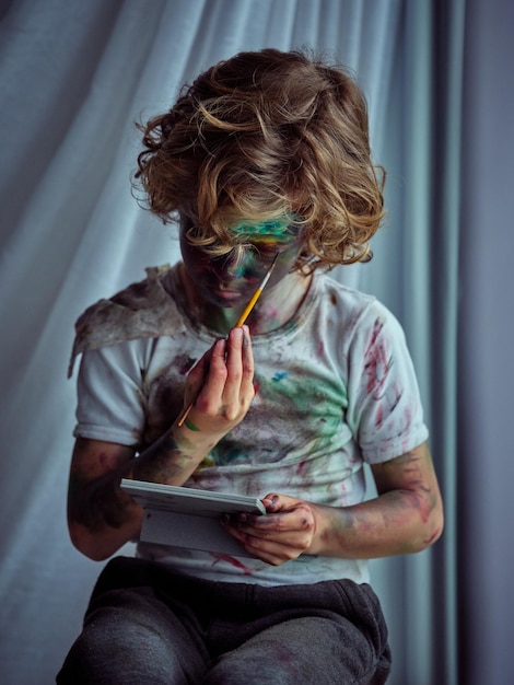 Enfant créatif portant un t-shirt sale avec des taches de peinture tenant un pinceau et se peignant à l'aide d'un miroir