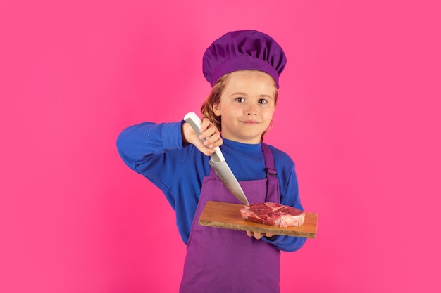 Enfant cuisinier tenir une planche à découper avec de la viande steak de boeuf et un couteau Cuisine enfants Chef enfant garçon faisant des aliments sains Portrait de petit enfant en toque isolé sur fond de studio