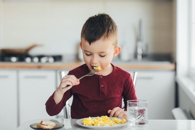 Un enfant dans l'après-midi sur une cuisine lumière blanche dans un pull bordeaux mange une omelette