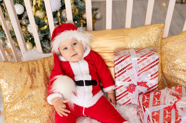 Un enfant dans un costume de père Noël avec un cadeau à l'arbre de Noël avec un décor doré est assis dans un berceau à la maison