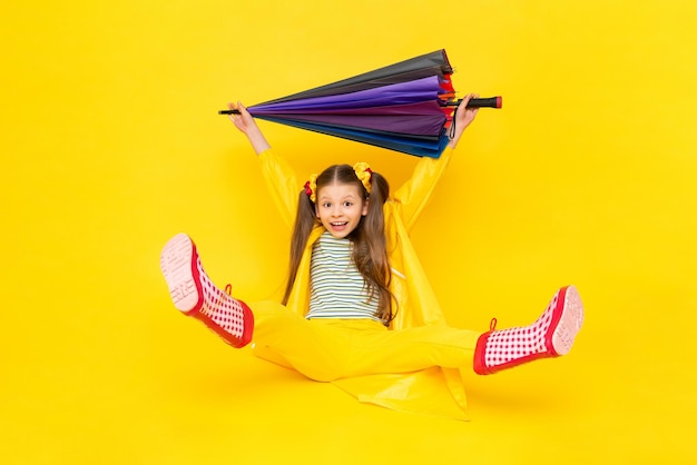 Un enfant dans un imperméable avec ses pieds dans des bottes rouges et tenant un parapluie multicolore au-dessus de sa tête Une belle petite fille sourit et est assise sur un fond jaune isolé Temps pluvieux