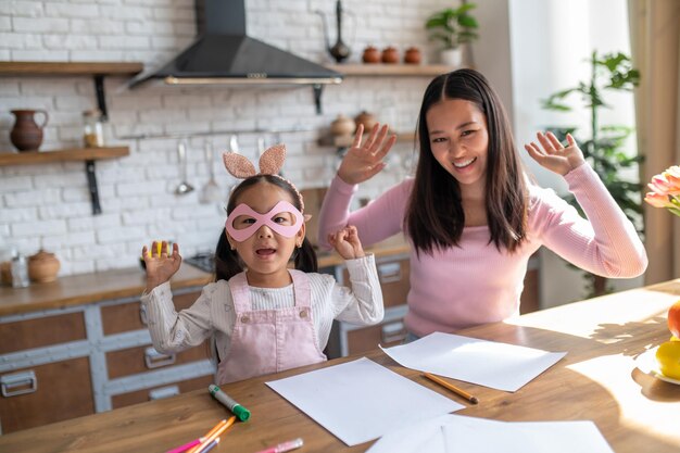 Enfant dans les lunettes en papier rose et sa joyeuse mère assise à la table s'amusant pendant l'enseignement à domicile