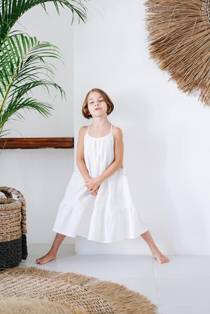 Un enfant dans une robe blanche les jambes étalées sur le fond d'un mur blanc d'une maison d'été