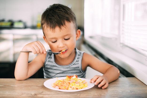 Un enfant dans un t-shirt dans la cuisine de manger une omelette avec des saucisses et des tomates avec une fourchette