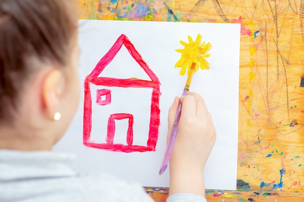 L'enfant dessine un soleil jaune avec une maison rouge.