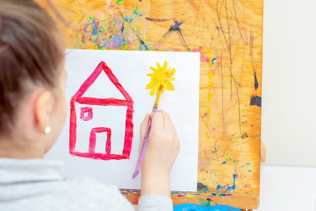 L'enfant dessine un soleil jaune avec une maison rouge.