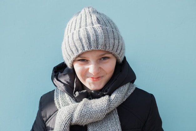 Enfant drôle en colère et mécontent prêt pour les vacances d'hiver. Garçon à la mode en bonnet gris d'hiver et écharpe debout contre le mur bleu.