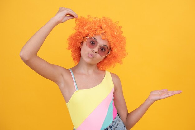 Enfant drôle en lunettes de soleil et maillot de bain portant des cheveux de perruque frisée orange sur fond jaune présentant la publicité de l'espace de copie du produit