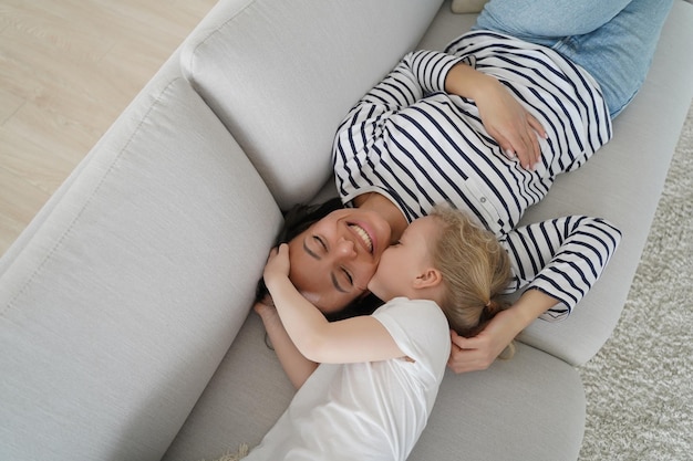 Enfant embrassant une mère heureuse. Jeune maman se reposant avec son petit enfant sur le canapé. La famille européenne joue et se détend ensemble. La femme et la fille s'amusent ensemble. Convivialité et amour.