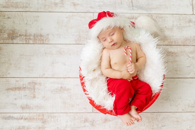 Un enfant endormi dans un costume de Père Noël se trouve avec un bonbon dans ses mains sur un fond en bois clair