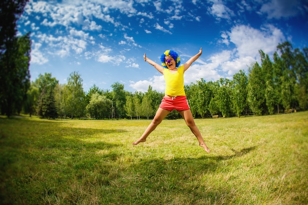 Enfant enfant fille avec la perruque bleue du parti clown drôle joyeux expression à bras ouverts et guirlandes saute dans le parc