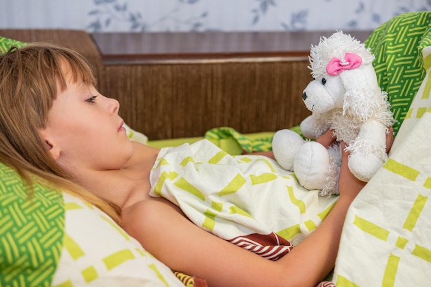 l'enfant est allongé dans son lit sous une couverture et tient un chien caniche jouet