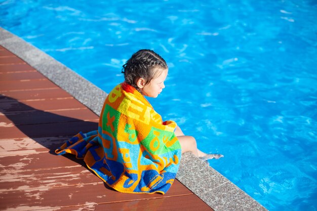 L'enfant est assis au bord de la piscine. Une petite fille avec des nattes tressées est assise au bord de la piscine et enveloppée dans...