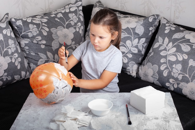 Un enfant est assis sur le canapé et fait une citrouille en papier mâché pour Halloween à partir d'un ballon