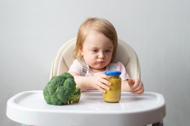 L'enfant est assis dans une chaise haute et hésite entre le brocoli frais et le ready-made en bocal