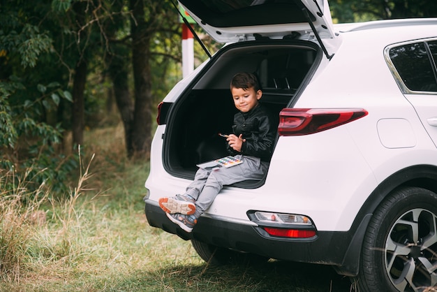 Un enfant est assis dans le coffre d'une voiture SUV ou d'un crossover avec des peintures à la gouache, une voiture dans la forêt d'automne