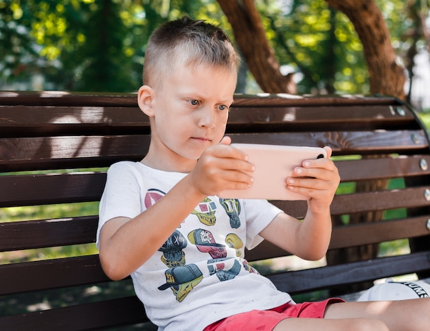 L'enfant Est Assis Dans Le Parc Sur Un Banc Avec Un Gadget. Les Enfants Utilisent Des Gadgets. Un Garçon Joue à Un Jeu Sur Un Téléphone Portable.