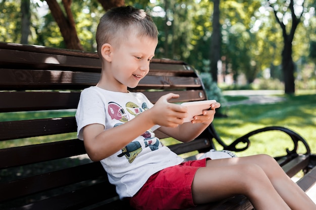 L'enfant est assis dans le parc sur un banc avec un gadget. Les enfants utilisent des gadgets. Un garçon joue à un jeu sur un téléphone portable.