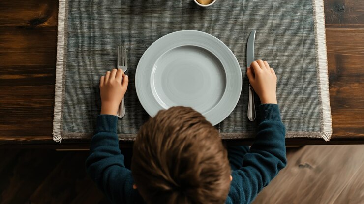 Un enfant est assis à une table à manger en bois, les mains de chaque côté d'une assiette grise, anticipant un repas.