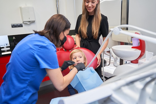 un enfant est en contrôle assis dans la chaise du dentiste avec sa mère en arrière-plan