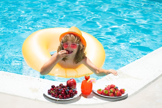 Enfant d'été au bord de la piscine mangeant des fruits et buvant un cocktail de limonade Concept de vacances pour enfants d'été Petit garçon se relaxant dans une piscine s'amusant pendant les vacances d'été