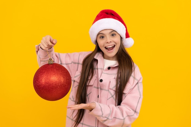 Un enfant étonné en chapeau de père noël tient une boule décorative de noël sur fond jaune, nouvel an.