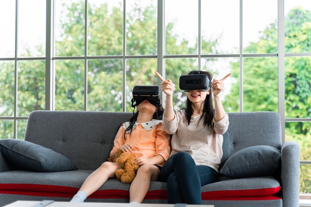 Enfant et femme avec casque de réalité virtuelle souriant assis sur un canapé à l'intérieur à la maison