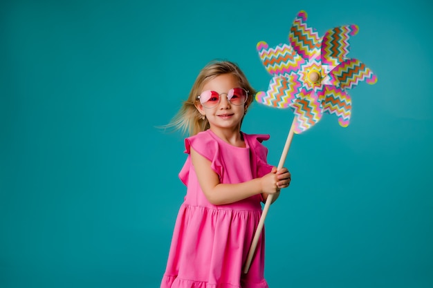 Enfant fille blonde souriant à lunettes de soleil dans une robe rose tenant un moulin à vent sur un fond bleu d'isoler