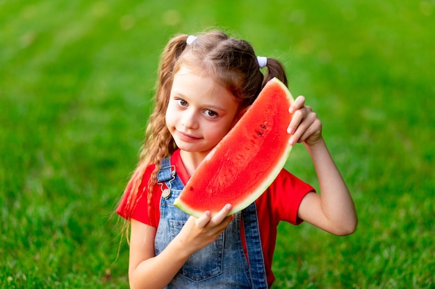 Une enfant fille en été sur la pelouse avec un morceau de pastèque sur l'herbe verte s'amuse et se réjouit de mordre l'espace pour le texte