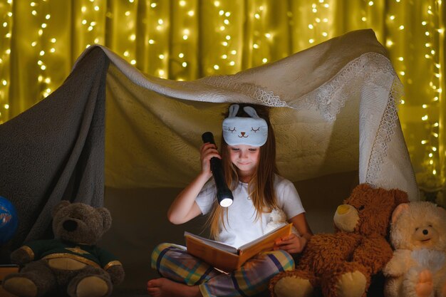 Enfant fille lisant avec livre et lampe de poche et ours en peluche dans la tente. avant d'aller au lit