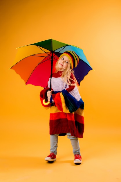 Enfant fille souriant dans un chandail multicolore lumineux et un chapeau avec un parapluie se dresse sur un espace jaune