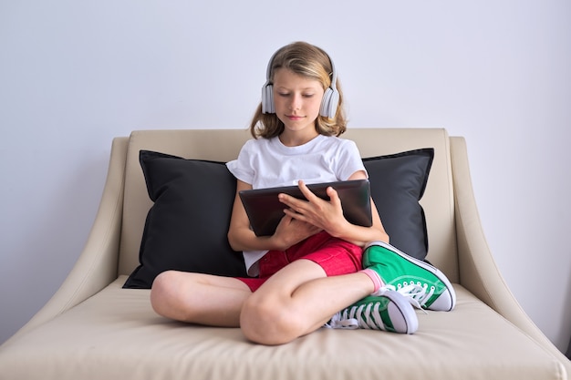 Enfant fille avec tablette numérique dans les écouteurs en regardant le moniteur assis sur le canapé à la maison