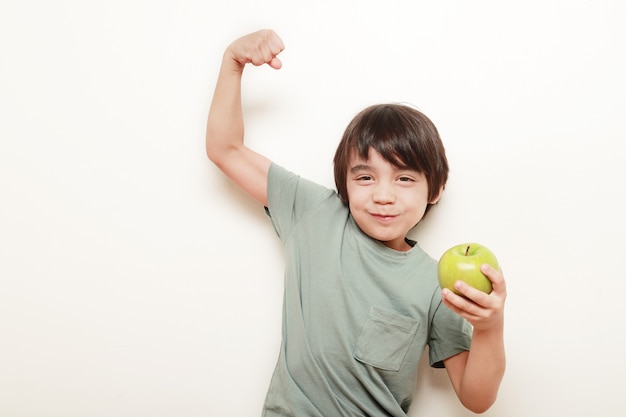 Enfant Sur Fond Blanc Mangeant La Pomme Verte Qu'il Tient Dans Une Main Et De L'autre Main, Il Nous Montre Son Biceps Indiquant La Force