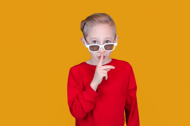 Un enfant gai dans des lunettes 3D pour enfants met son index sur ses lèvres et fait un geste calme