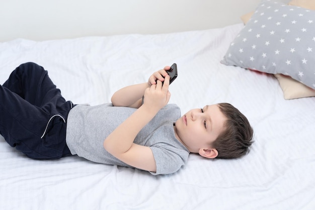 Enfant garçon allongé sur le lit avec un smartphone jouant à des jeux ou étudiant quelque chose dessus Concept social et technologique