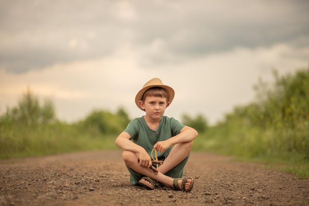 Photo enfant garçon dans un chapeau avec une fronde dans ses mains est assis sur la route