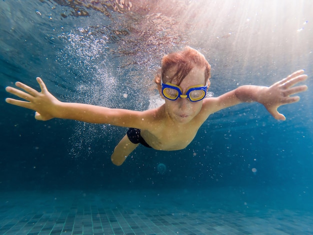 Un enfant garçon nage sous l'eau dans une piscine, souriant et retenant son souffle, avec des lunettes de natation