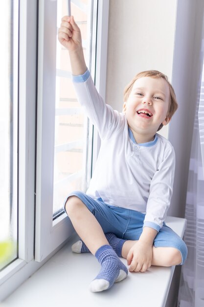 Un enfant garçon regarde par la fenêtre assis sur le rebord de la fenêtre de la maison.