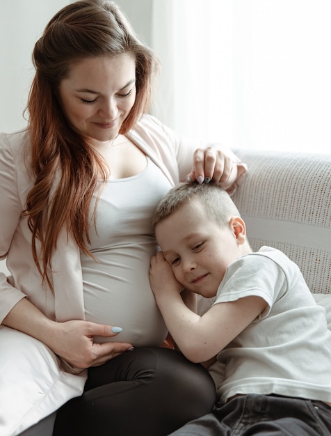 Enfant garçon serrant le ventre de sa mère enceinte sur le canapé à la maison.