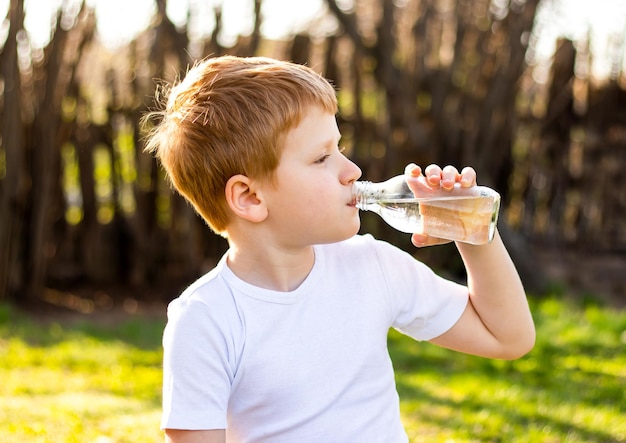 enfant garçon en T-shirt blanc buvant de l'eau froide propre à partir d'une bouteille assis sur l'herbe dans la nature