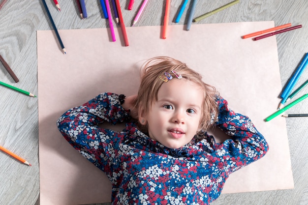 Enfant gisant sur le sol sur du papier près de crayons de couleur Petite fille peignant, dessin Concept de créativité